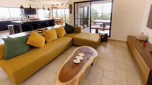 Una sala moderna con un sofá grande, una mesa de centro de madera y una zona de cocina con una barra y comedor visibles a través de puertas de vidrio.