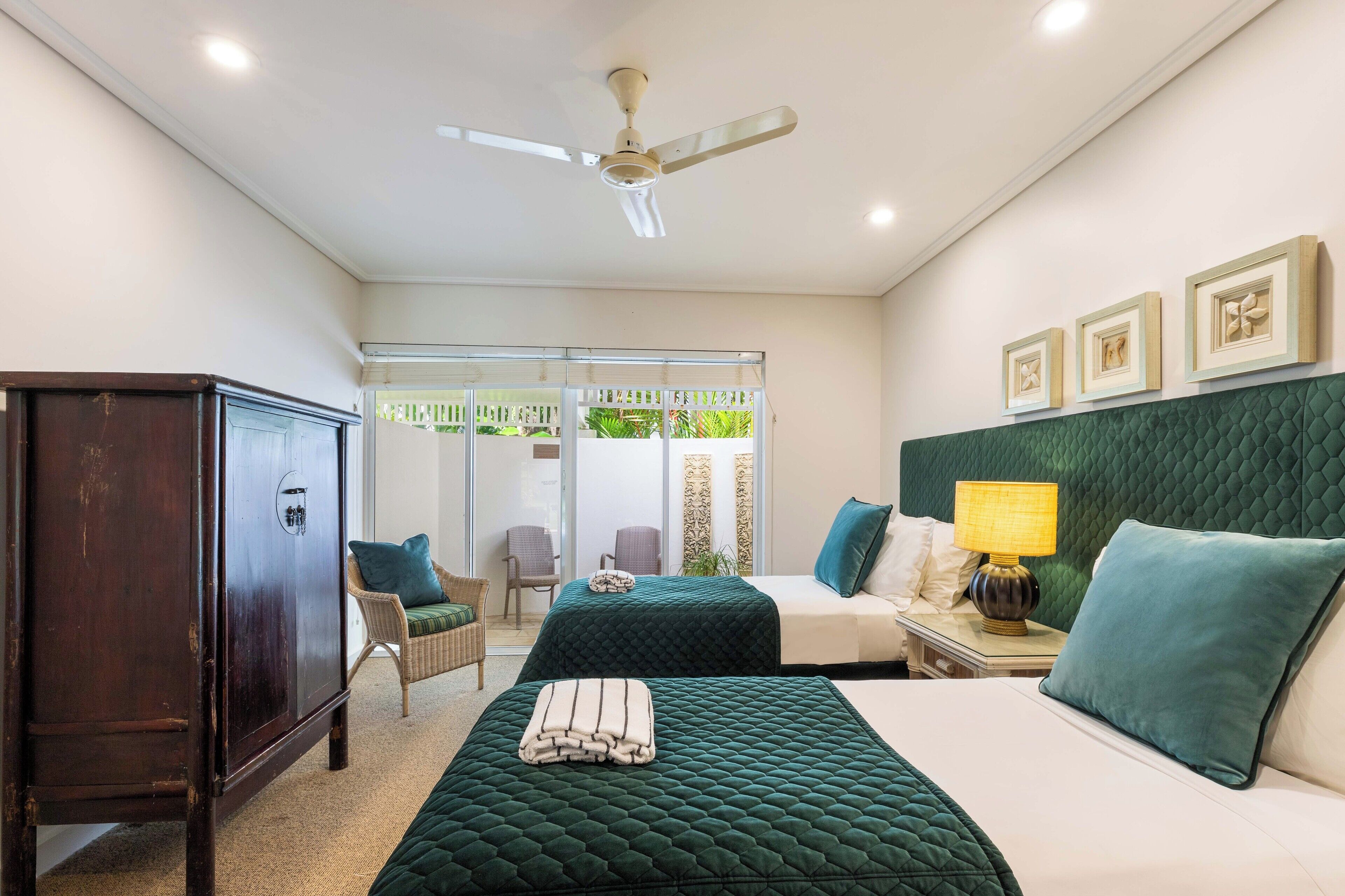 Quarto de hotel com duas camas, um ventilador de teto, um guarda-roupa, uma cadeira, uma mesa de cabeceira com abajur e vista para o exterior por uma porta de vidro.