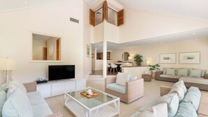 A spacious living area with a glass coffee table, multiple seating options including a sofa and armchairs, a flat-screen TV, and a dining area with a table and chairs.