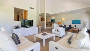 Una sala de estar moderna con muebles blancos, un televisor de pantalla plana y un comedor al fondo.