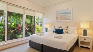 Un dormitorio con una cama grande, una mesita de noche, una lámpara y vistas a la vegetación a través de amplias ventanas.