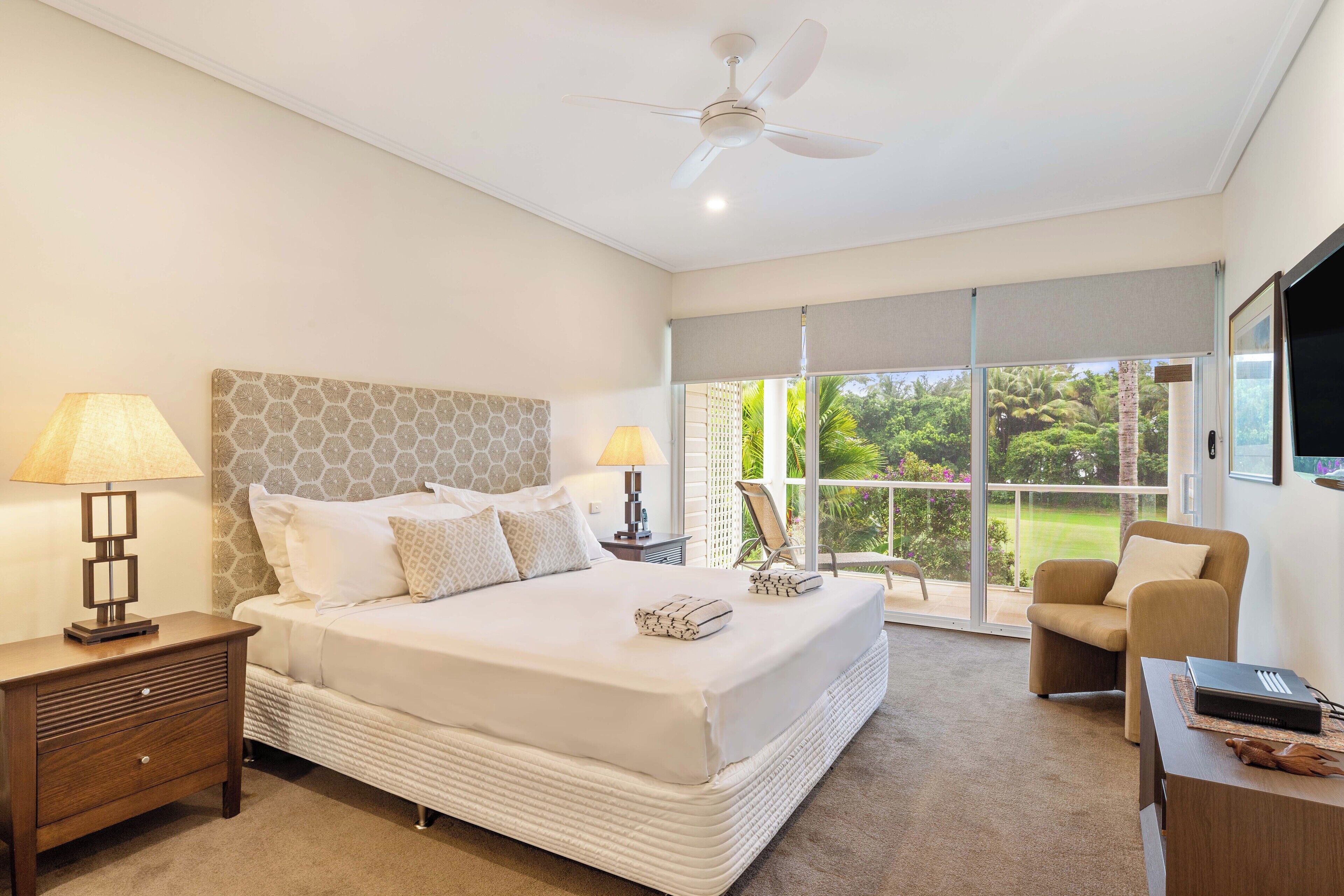 A bedroom with a large bed, two bedside lamps, a nightstand, a chair, a desk, and a ceiling fan.