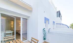 Une chambre d’hôtel moderne dotée d’une porte-fenêtre coulissante donnant sur un balcon, d’un banc en bois et d’un tuyau d’arrosage vert.