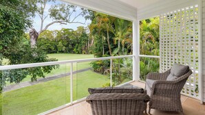 A balcony with a wicker chair and a small table, overlooking a well-maintained garden with various trees and a clear path. - Mirage Resort Villa 420 with access to Sheraton Facilities (Port Douglas)