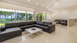 Uma sala de estar moderna com sofá seccional, mesa de centro de vidro e amplas janelas com vista para áreas verdes.