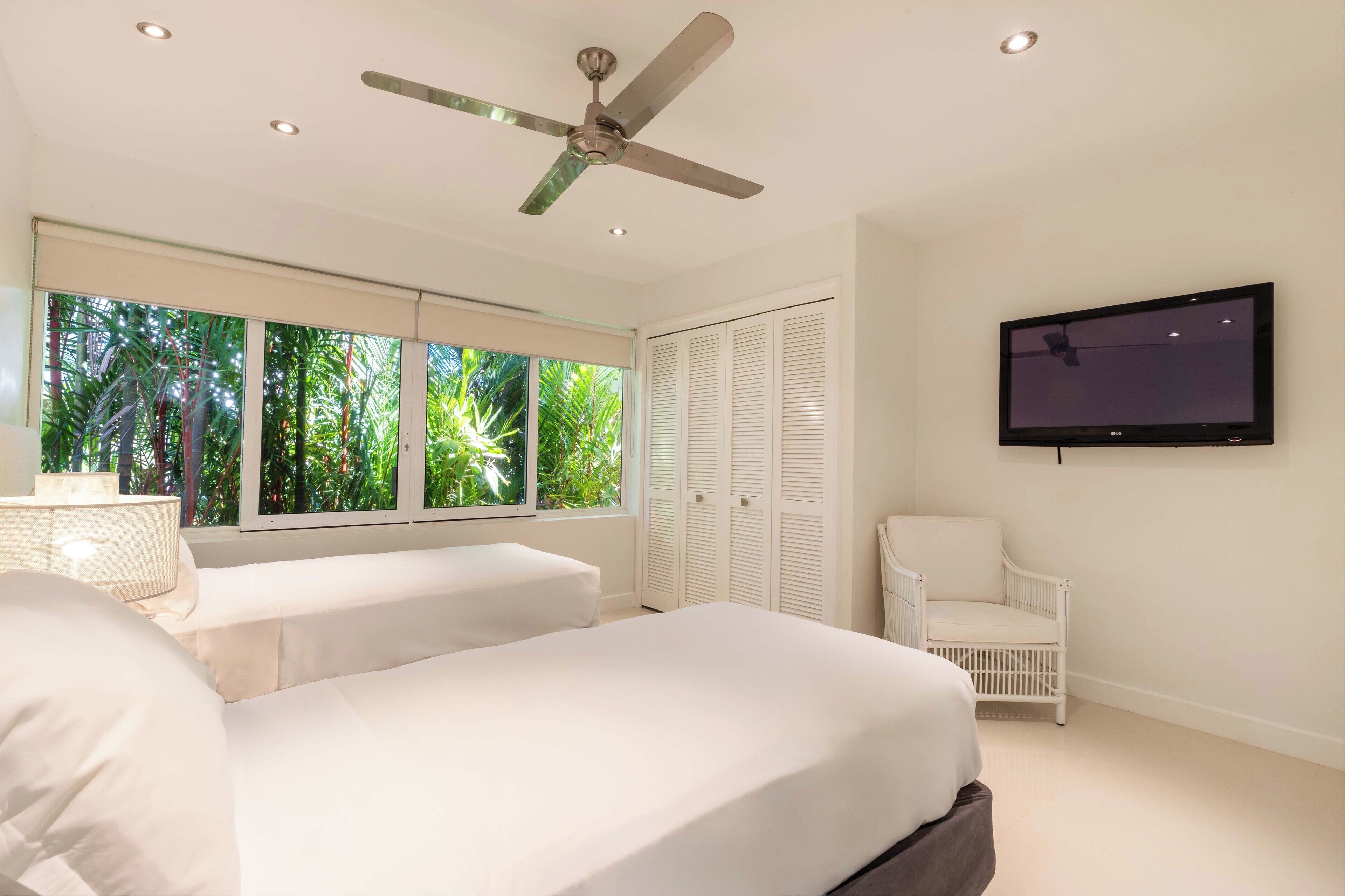 A bedroom with a bed, a chair, a TV, a ceiling fan, and a window with a view of greenery.