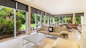 Un amplio salón con un gran ventanal corredera que da a un jardín exuberante. Hay varias opciones de asientos, incluyendo un sofá y sillones.