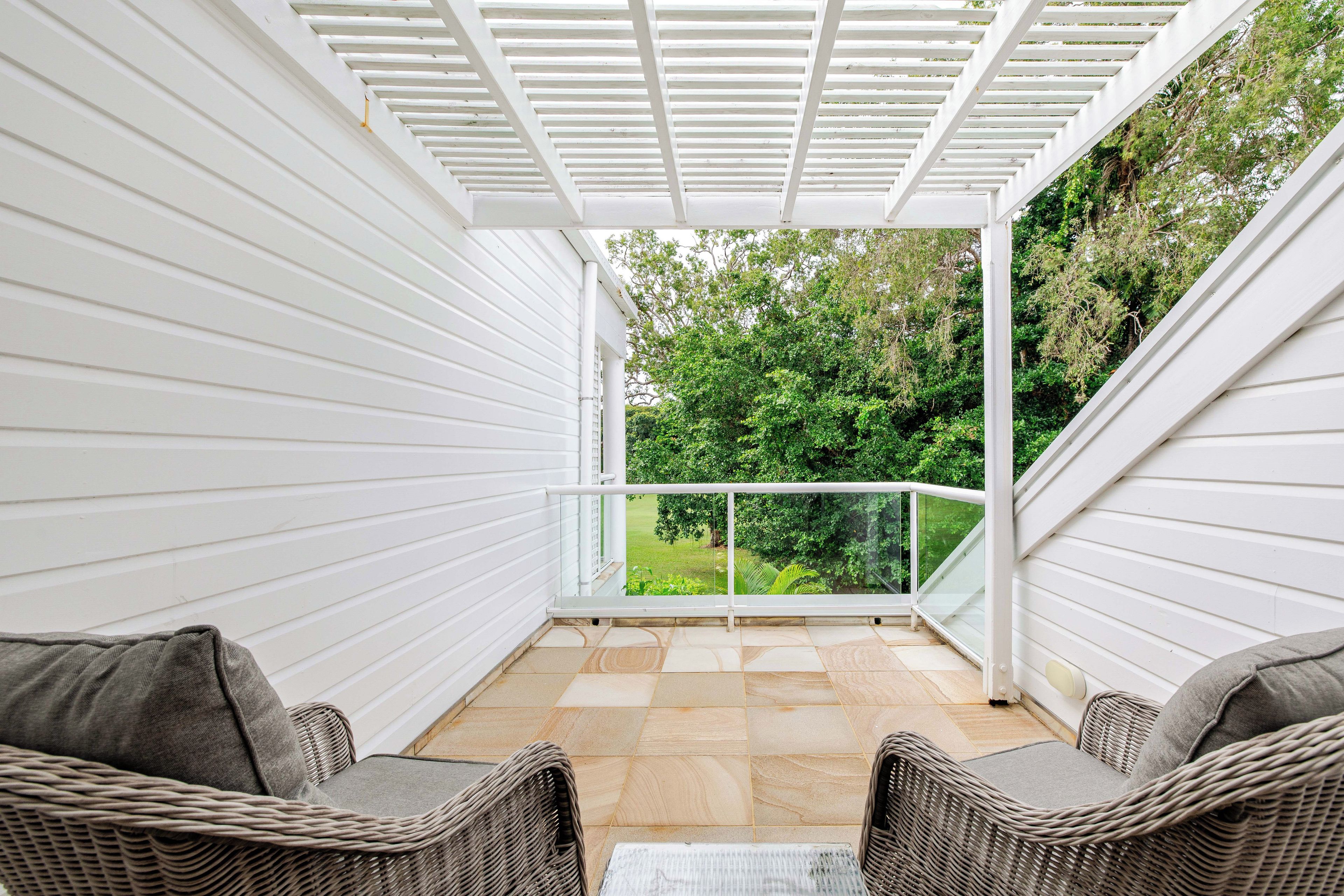 Une terrasse couverte avec deux fauteuils en osier, une balustrade en verre et une vue sur la verdure.