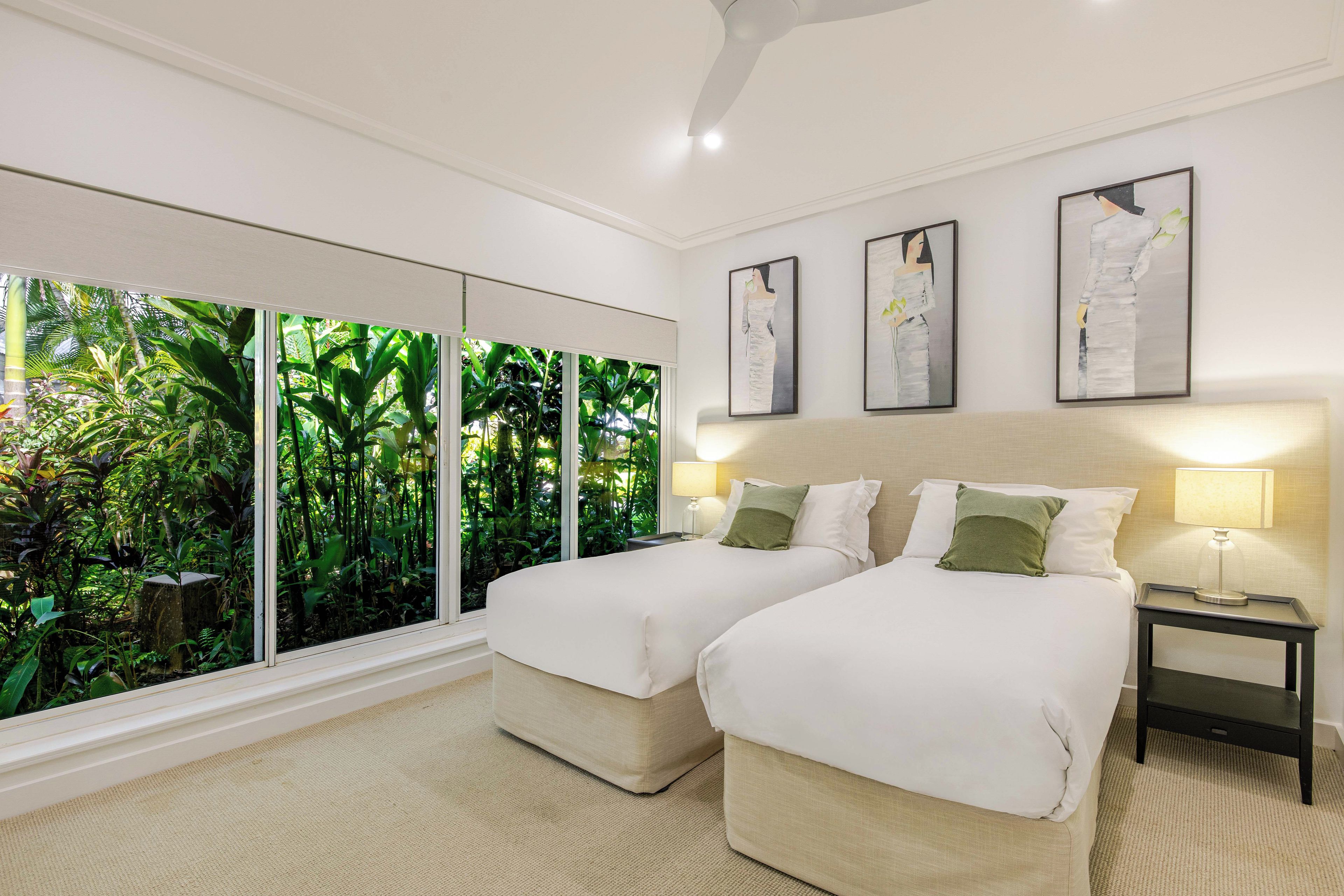 A hotel room with two beds, a large window with a view of greenery, and three framed artworks on the wall.