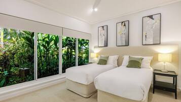 A hotel room with two beds, a large window with a view of greenery, and three framed artworks on the wall.