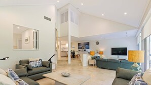 Uma sala de estar moderna com um sofá seccional, uma mesa de centro e uma TV. Ao fundo, há uma cozinha com uma mesa de jantar e cadeiras.