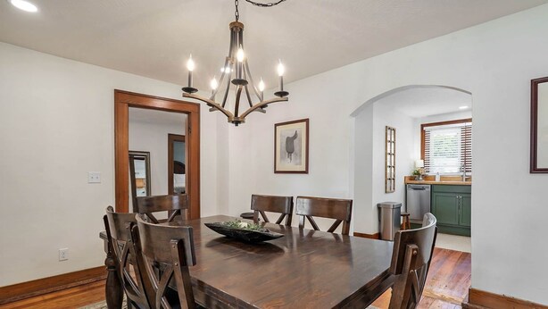 A dining room with a wooden table, six chairs, a chandelier, and a kitchen in the background.