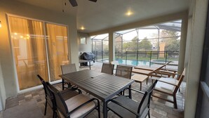 An outdoor patio with a table and chairs, a covered grill, and a view of a swimming pool. - 8909 Disney Retreat – 6BR Home with Pool & Comfort (Kissimmee)
