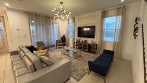 A modern living room with a sofa, chairs, a coffee table, a TV, and a blue ottoman. - 8909 Disney Retreat – 6BR Home with Pool & Comfort (Kissimmee)