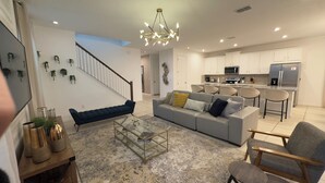 A modern living room with a sofa, coffee table, and chairs. A staircase leads to another level. The kitchen features white cabinets, a refrigerator, and a microwave. - 8909 Disney Retreat – 6BR Home with Pool & Comfort (Kissimmee)