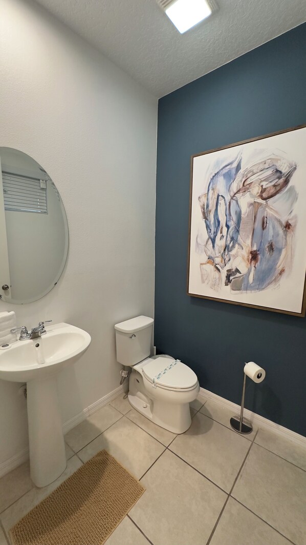 A bathroom with a white sink, a toilet, a blue accent wall, and a painting. - 8909 Disney Retreat – 6BR Home with Pool & Comfort (Kissimmee)