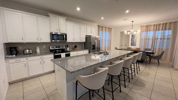 A modern kitchen with white cabinets, stainless steel appliances, a granite countertop, and a bar area with stools. - 8909 Disney Retreat – 6BR Home with Pool & Comfort (Kissimmee)