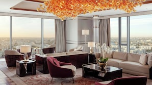 A modern hotel room with a large city view, a plush sofa, a red armchair, a coffee table, and a decorative chandelier. - JW Marriott Hotel Tashkent (Tashkent)