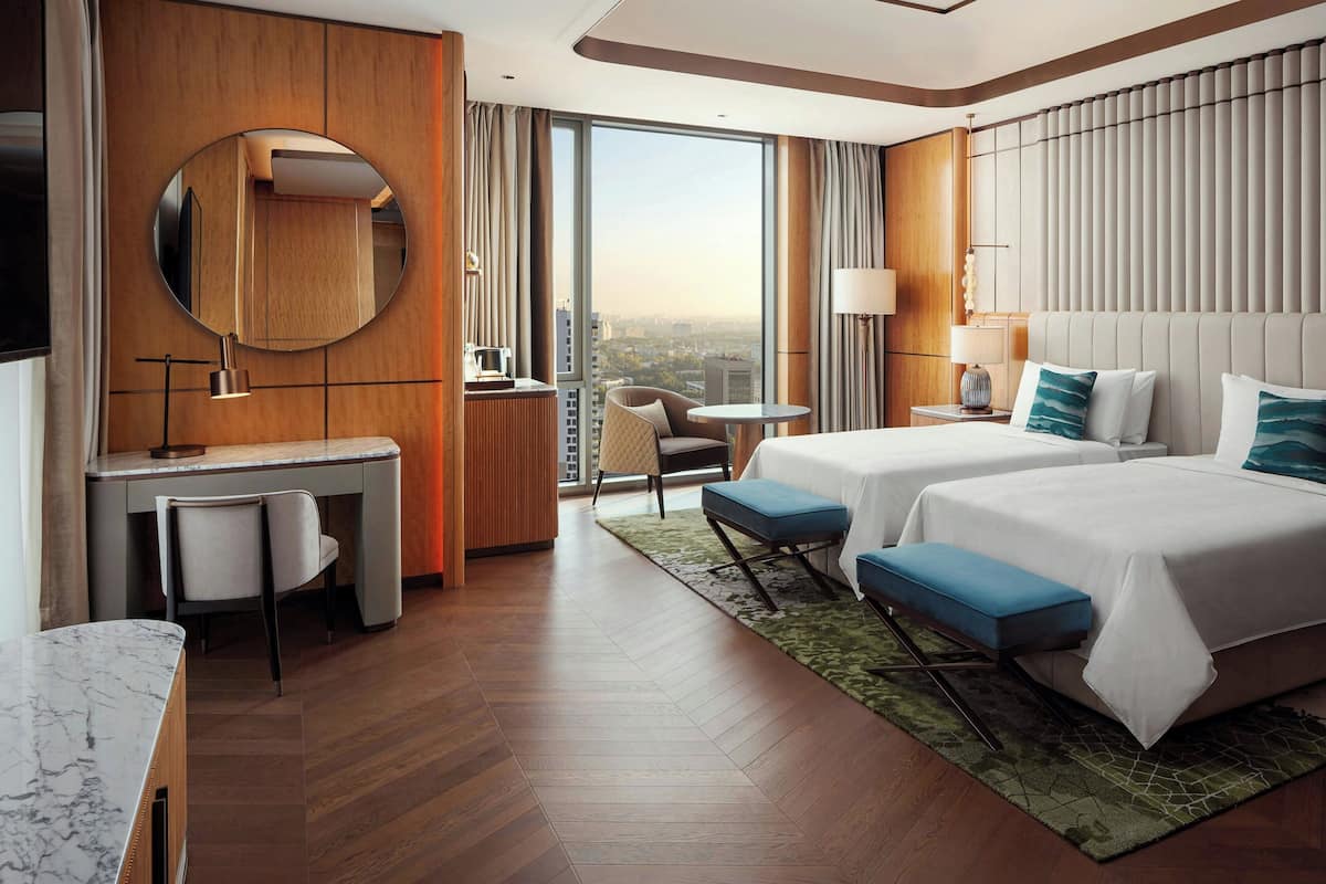 Habitación de hotel con dos camas, un escritorio, una silla, un espejo redondo y vistas a la ciudad.