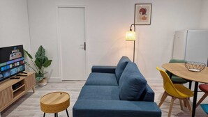Ett modernt vardagsrum med en blå soffa, ett soffbord i trä, en liten krukväxt, en tv på ett träställ, ett matbord med färgglada stolar och en vägglampa.