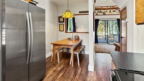 Eine kompakte Küche mit Edelstahlkühlschrank, einem Holzespeisetisch und einer Bank, einer Pendelleuchte sowie Blick auf eine Terrasse durch eine Glasdoor.