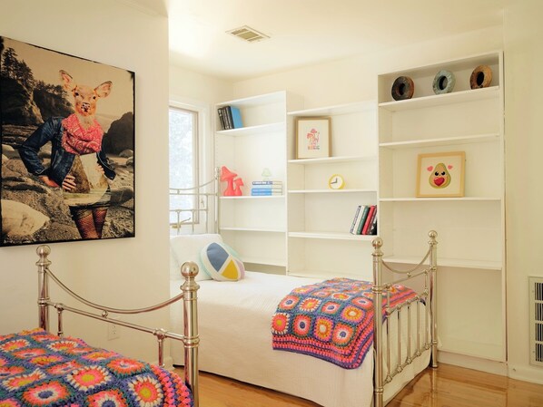 Un dormitorio con estructura de cama metálica, una colcha colorida, una estantería y un cuadro en la pared.