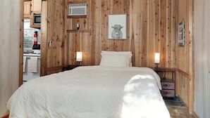 Um quarto com painéis de madeira, uma cama, abajures, um quadro emoldurado e uma kitchenette ao fundo.