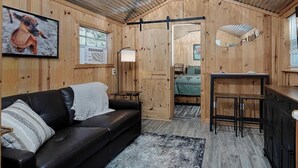 A cozy cabin interior with a wooden interior, a black leather sofa, a small dining area, and a bed with a green blanket. - Pixie's Porch Tiny Home (Centerville)
