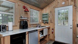 A compact kitchen with wooden walls, a white door, a sink, a coffee maker, a toaster, and a small refrigerator. - Pixie's Porch Tiny Home (Centerville)
