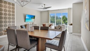 A modern living room with a wooden dining table, grey upholstered chairs, a ceiling fan, and a large window with a view of greenery.