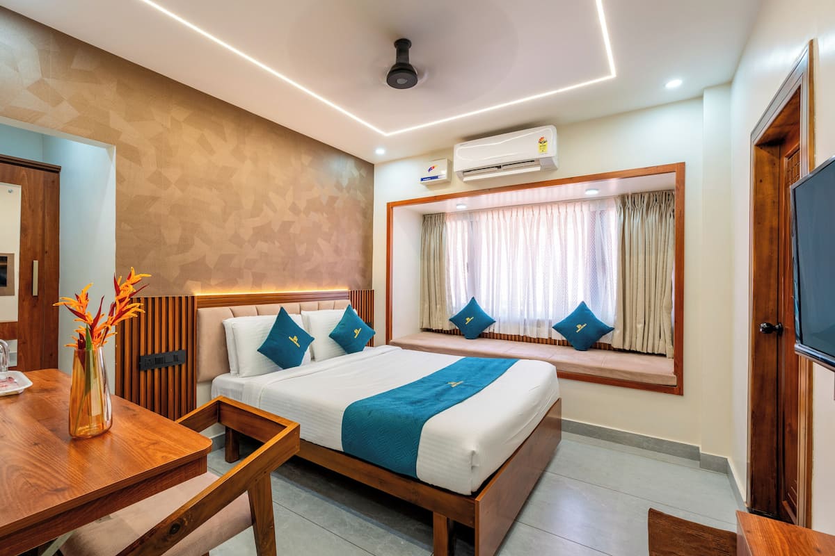 A hotel room with a bed, a wooden desk, a vase with flowers, an air conditioning unit, and a window with curtains.