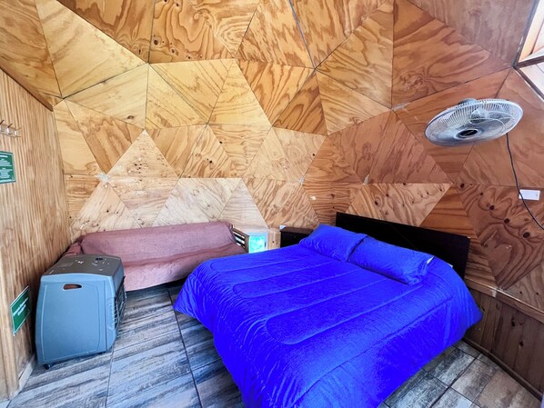 Una habitación con techo de madera con diseño geométrico, una cama con cubrecama azul, una mesita pequeña y un ventilador.