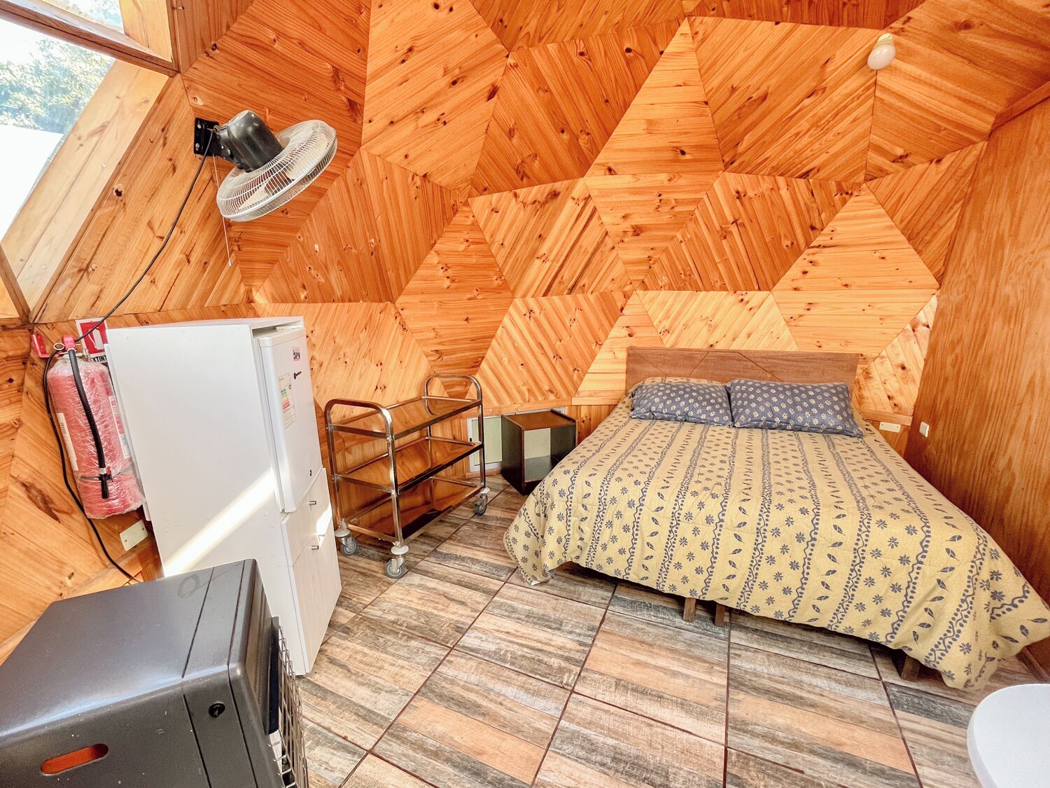 Quarto compacto em formato triangular, com uma cama, uma mesa pequena, um ventilador e um frigorífico.