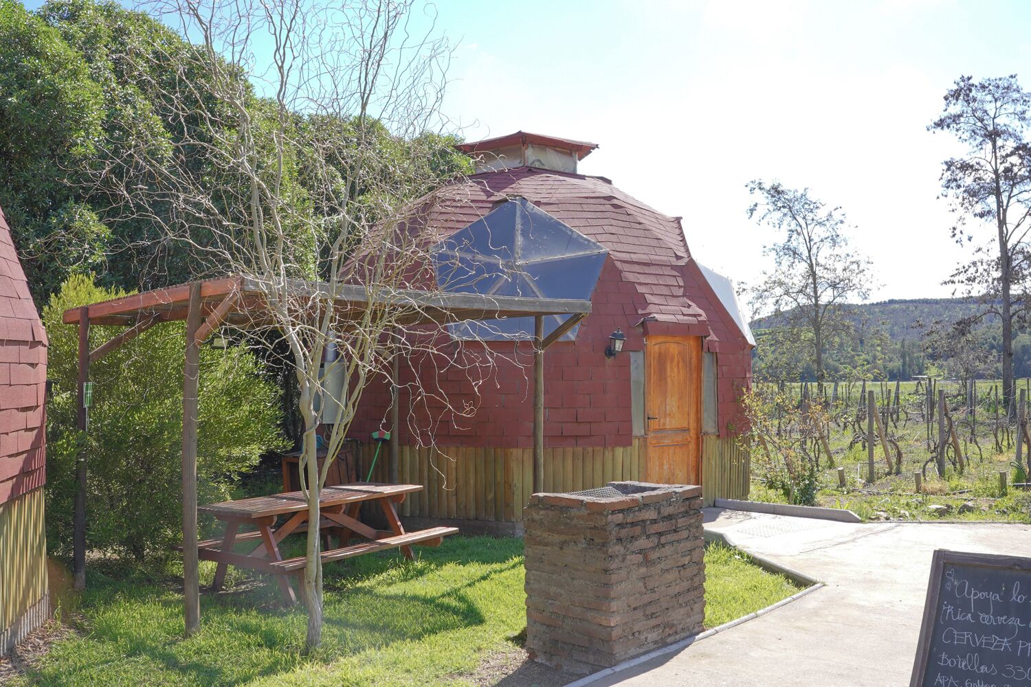Un área de estar al aire libre de estilo rústico con una mesa de madera y bancos, una estructura de ladrillo con techo inclinado y un cartel.