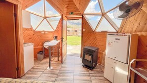 A cozy cabin interior with a wooden ceiling, a small kitchenette, a dining area, and large windows offering a view of the outdoors. - DOmos Ocoa (Hijuelas)