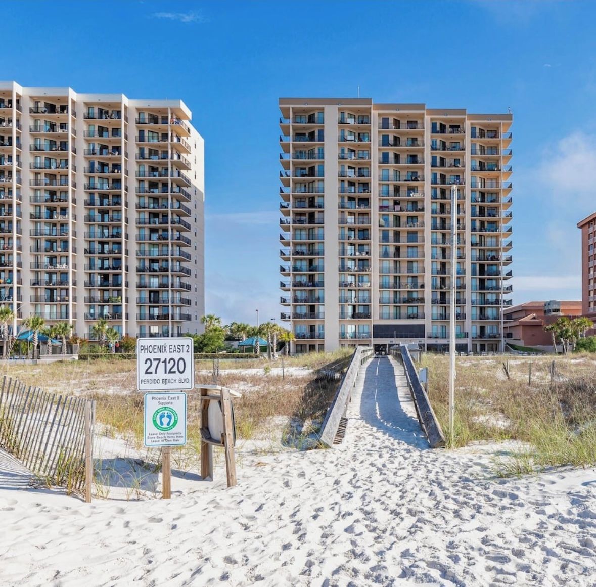 Un complesso di condomini fronte spiaggia con diversi edifici a grattacielo, cielo sereno e un vialetto sabbioso che conduce alla spiaggia.