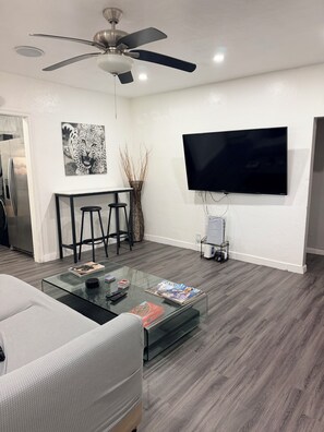 A modern living room with a flat-screen TV, a ceiling fan, a glass coffee table with magazines, a white sofa, and a kitchen area with a refrigerator.