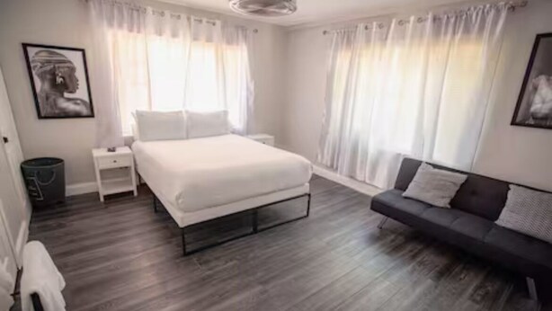 A bedroom with a bed, a bedside table, a nightstand, a sofa, and a large window with curtains.
