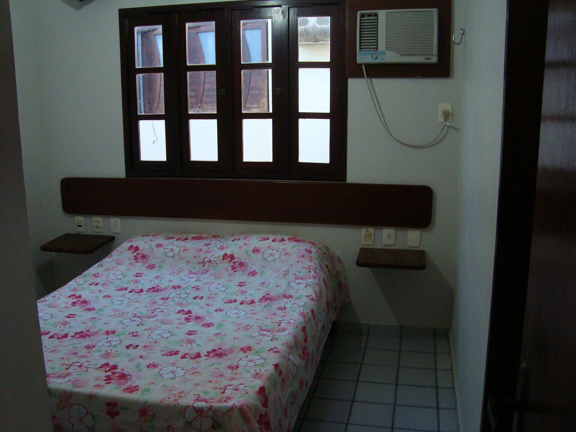 A small bedroom with a bed, a window, a wall-mounted air conditioner, and a bedside table.