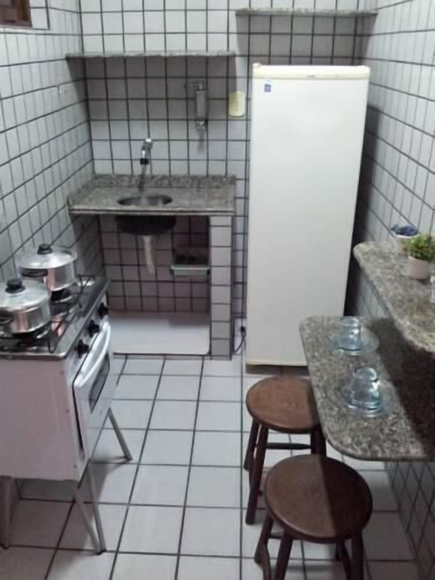 Cocina compacta con estufa a gas, lavaplatos y refrigerador blanco. Hay dos taburetes y una mesa pequeña con una planta.