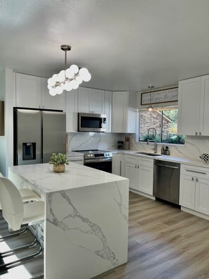 Une cuisine moderne avec des meubles blancs, un îlot en marbre, des appareils électroménagers en acier inoxydable et une vue sur un mur de briques.
