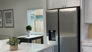 A modern kitchen with white cabinetry, a stainless steel refrigerator, a marble countertop, and a pendant light fixture. - The Woodlands Retreat Getaway & Theater Luxury (The Woodlands)
