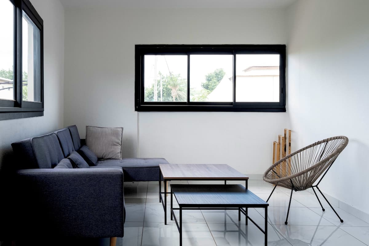 Ein modernes Wohnzimmer mit einer Eckcouch, einem kleinen Tisch, einem Korbstuhl und einem großen Fenster mit Blick auf Grünflächen.
