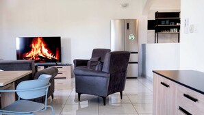 Una sala moderna con televisor de pantalla plana, chimenea, comedor con mesa y sillas, y cocina con refrigerador y alacenas.