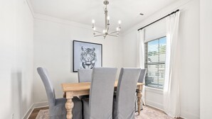 A dining area with a wooden table, grey upholstered chairs, a chandelier, a tiger artwork, and a window with blinds. - Tiger’s Tail Trail 1245~Aubie's Reserve (Auburn)