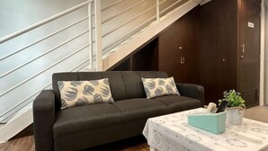 A dark-colored sofa with patterned cushions, a small table with a patterned tablecloth, and a staircase with white railings. - The CEO SuiteZ1 By Homey Planet (Bayan Lepas)