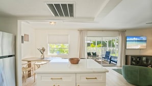 A modern kitchen with a central island, a dining area with a table and chairs, a TV mounted on the wall, and a view of a balcony with outdoor seating. - SHELL-HAUS | Venice Beach House | 5-min to Gulf (Venice)