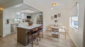 A modern kitchen with white cabinetry, a central island with stools, and a dining area with a round table and chairs. - SHELL-HAUS | Venice Beach House | 5-min to Gulf (Venice)