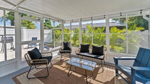 A covered patio with wicker furniture, a glass table, and large windows offering a view of greenery. - SHELL-HAUS | Venice Beach House | 5-min to Gulf (Venice)