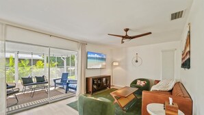 A spacious living room with a sliding glass door leading to a balcony, a ceiling fan, a television, and a green armchair. - SHELL-HAUS | Venice Beach House | 5-min to Gulf (Venice)
